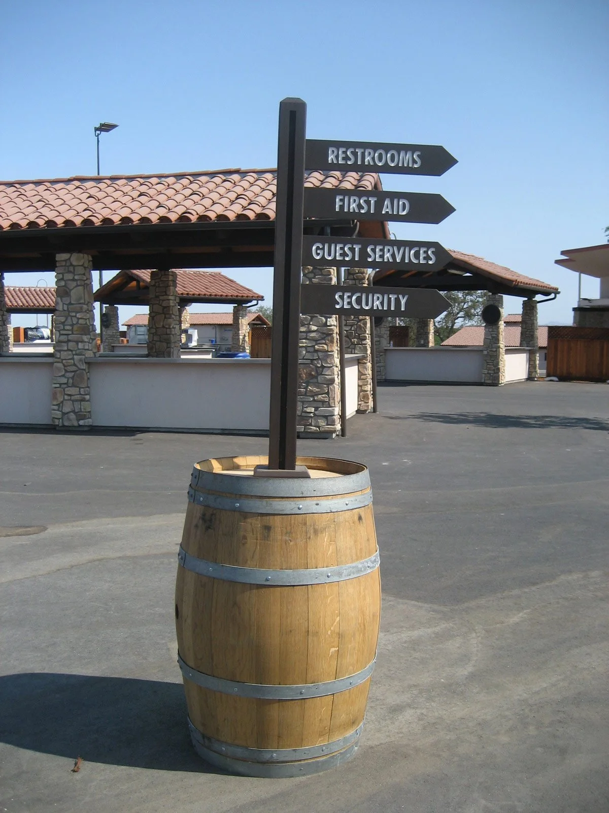A wooden barrel repurposed as a base for a signpost with four black directional signs indicating restrooms, first aid, guest services, and security, in an outdoor area with stone pillars, tiled roofs, and paved ground.