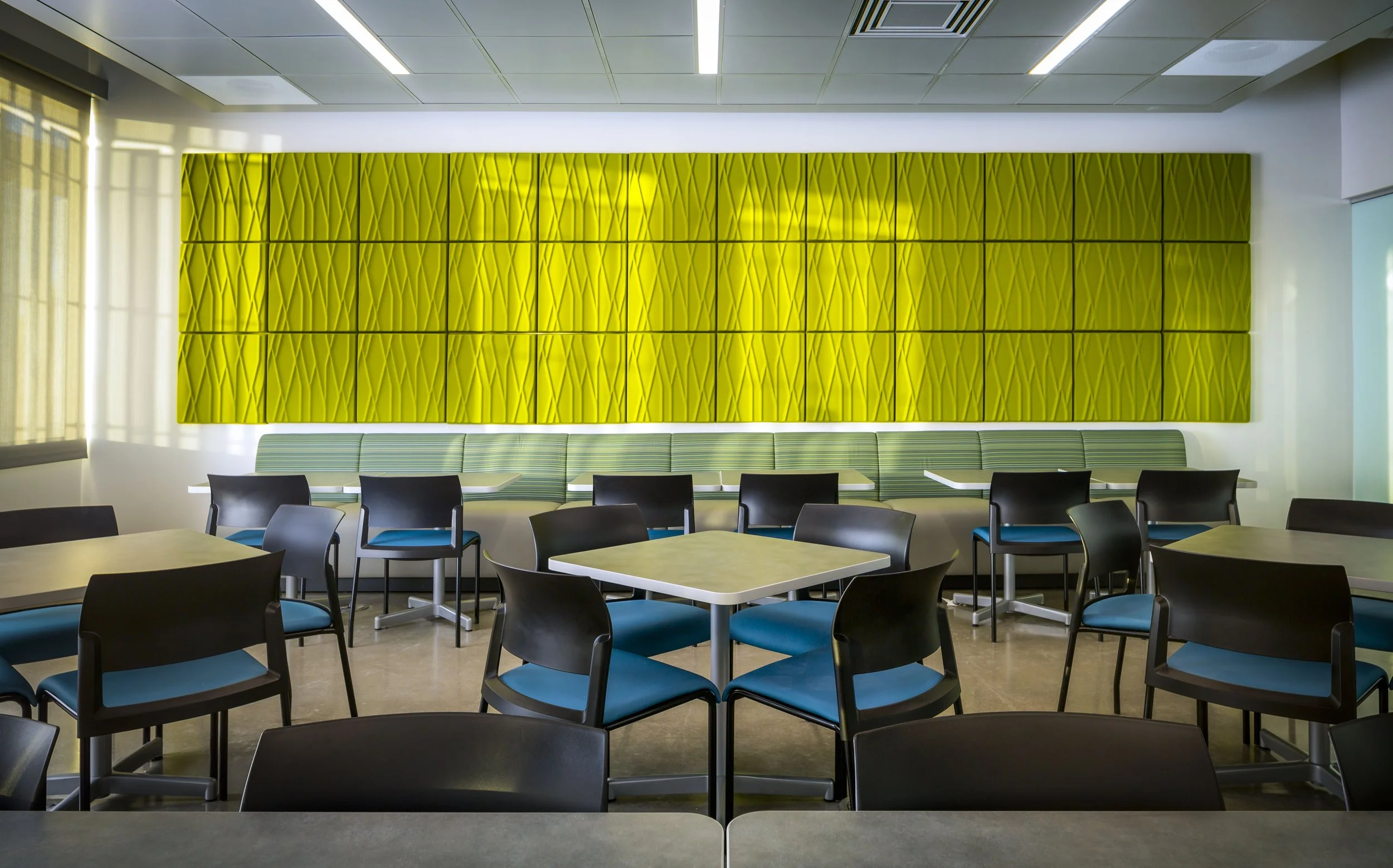 Empty dining area with black and blue chairs around square and round tables, a long green cushioned bench against a bright yellow decorative wall, and windows letting in natural light.