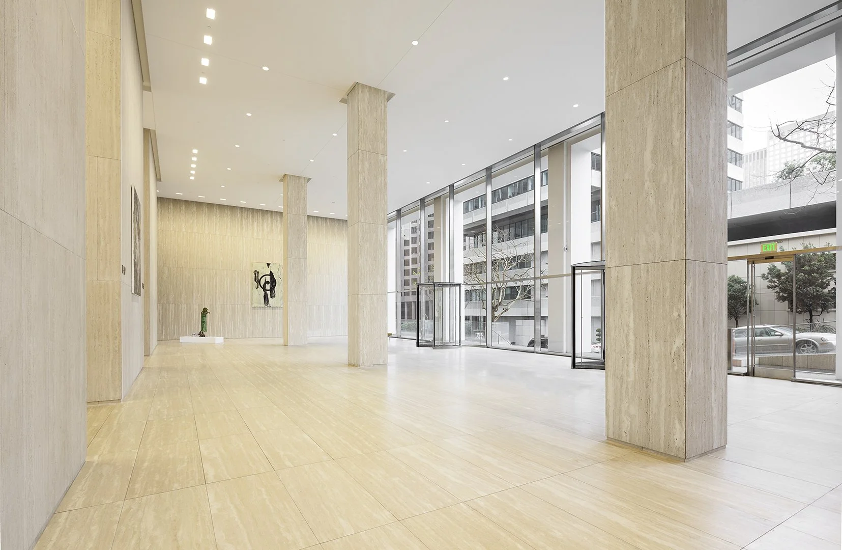 Modern art gallery with large windows, beige tiled walls and floor, sculptures, and artwork displayed on the walls.