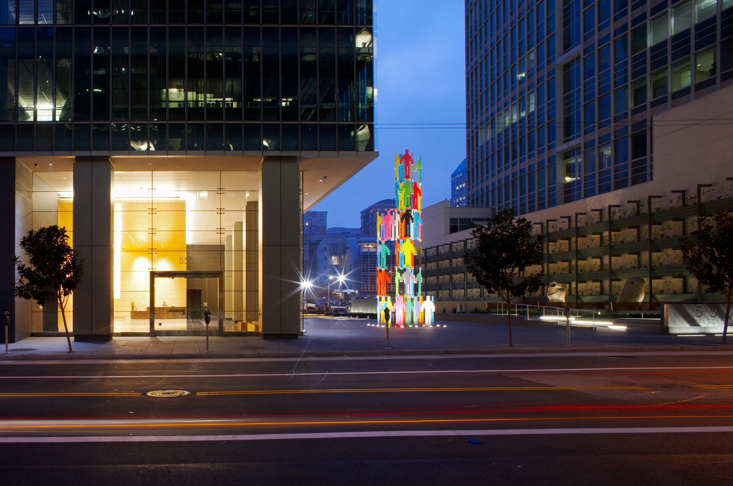 Multicolored illuminated sculpture of human figures in an urban environment at dusk, surrounded by modern buildings and trees.