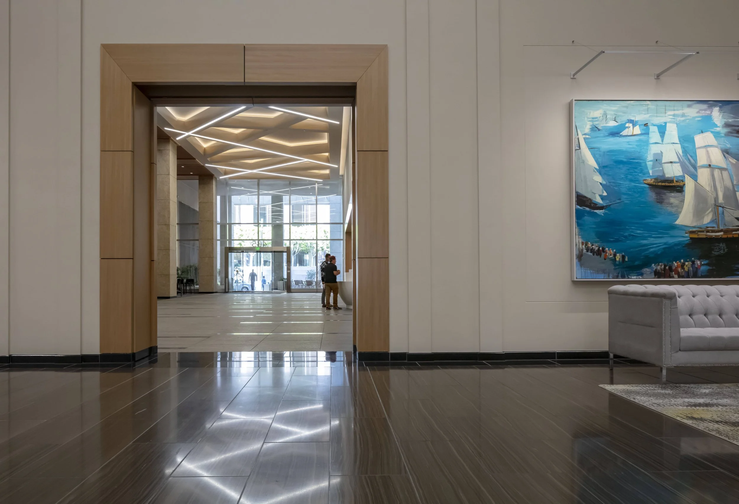 Interior of a modern building with a hallway leading to an entrance, featuring art on the wall, a gray tufted sofa, and a large digital painting of sailboats on the water.