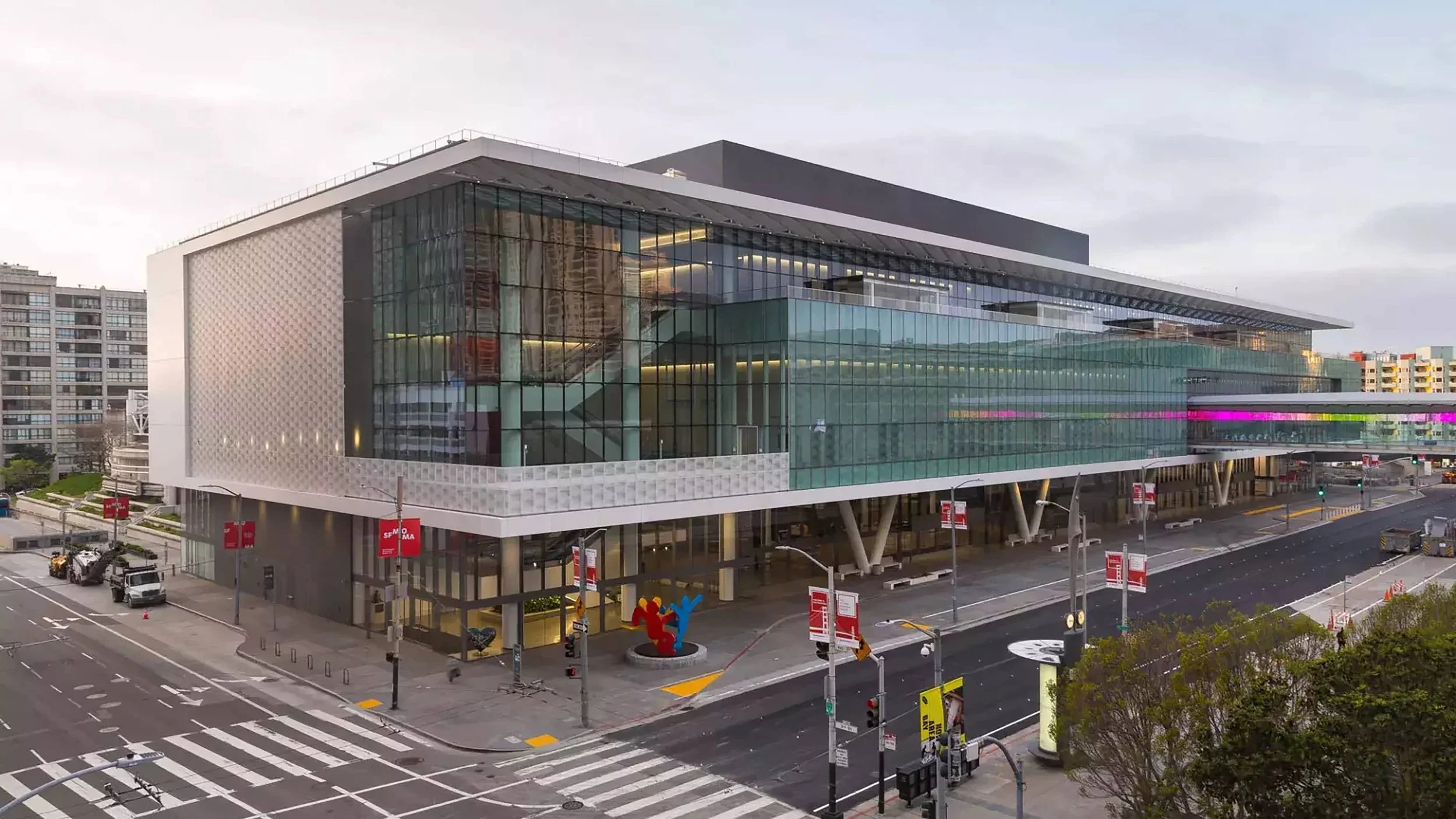 A modern multi-story glass building with a white structural frame located in an urban area. The building features large glass windows, a colorful sculpture near the entrance, and roadways with traffic signals and crosswalks in the foreground. The scene appears to be captured during early evening or late afternoon with overcast skies.