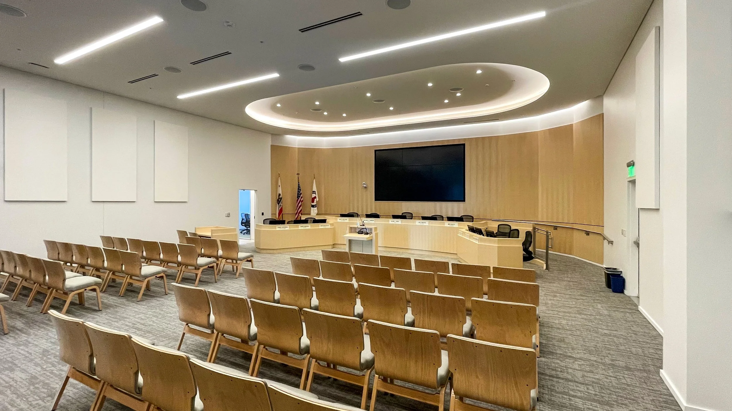 Empty modern conference room with wooden chairs arranged facing a stage with flags, a large screen, and a curved wood-paneled wall.
