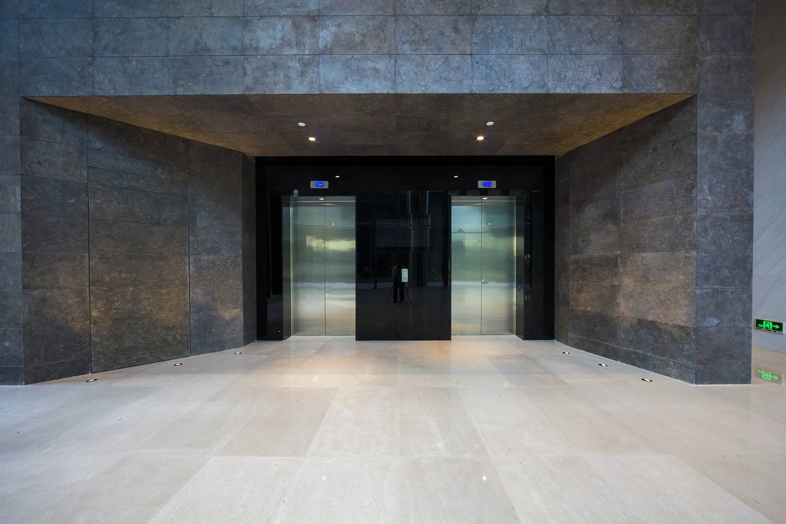 Modern building entrance with two glass elevators, dark stone walls, and a beige tiled floor.