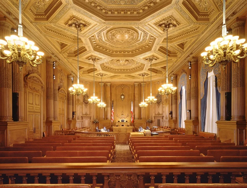 A grand courtroom with ornate wood paneling, chandeliers, and large windows with blue curtains, set for a formal hearing or trial.