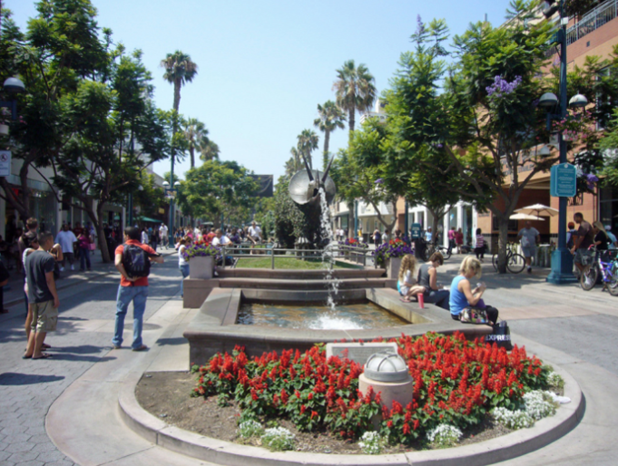 Outdoor shopping area with a fountain, surrounded by colorful flowers, trees, and people walking and sitting.