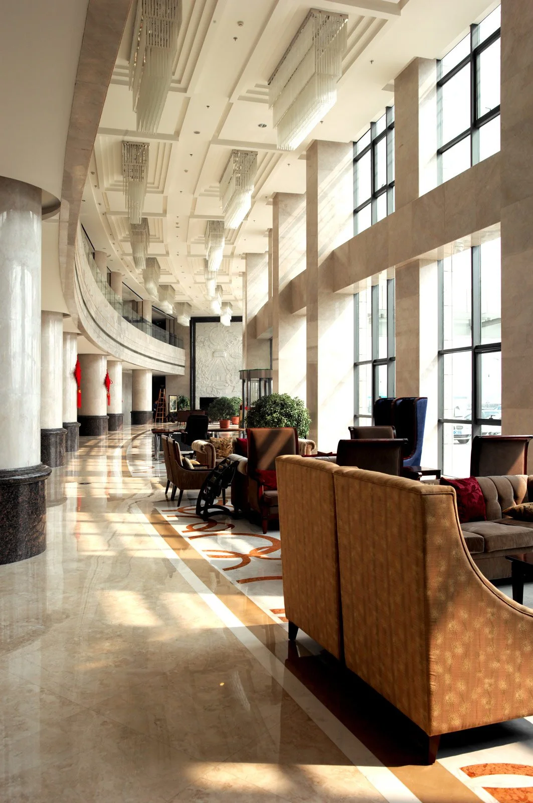 A spacious hotel lobby with high ceilings, large windows allowing natural light, elegant chandeliers, and plush armchairs and sofas arranged for guests, with potted plants and a polished marble floor.
