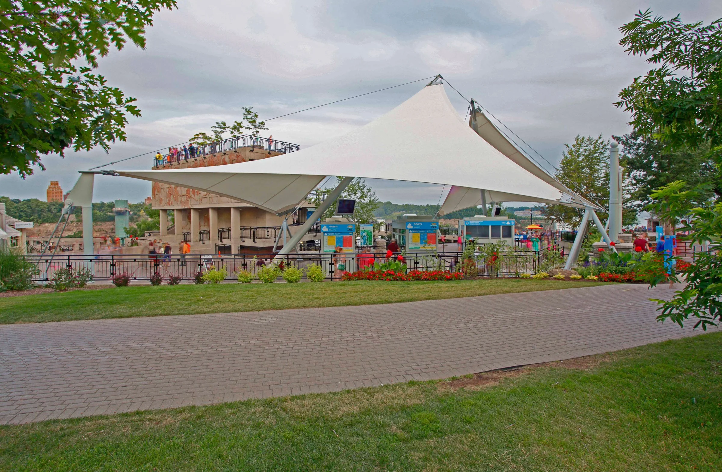 View of a modern amusement park ride structure with a large white canopy, ticket booths, and people walking around on a cloudy day.