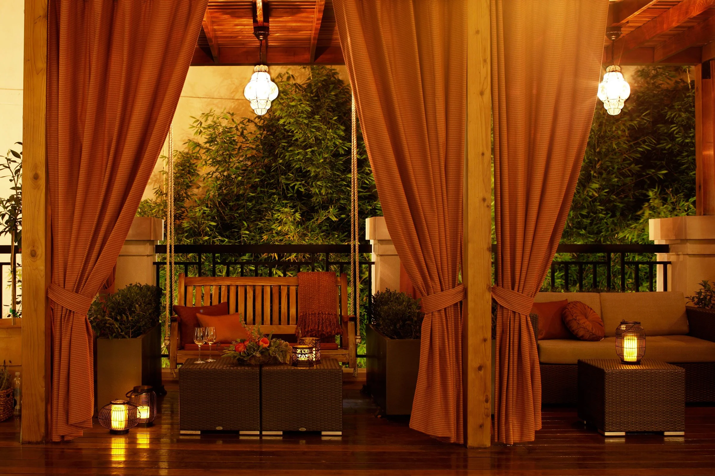 Outdoor seating area under a wooden pergola with curtains, candles, plants, a swing, and a sofa, illuminated by warm lighting.