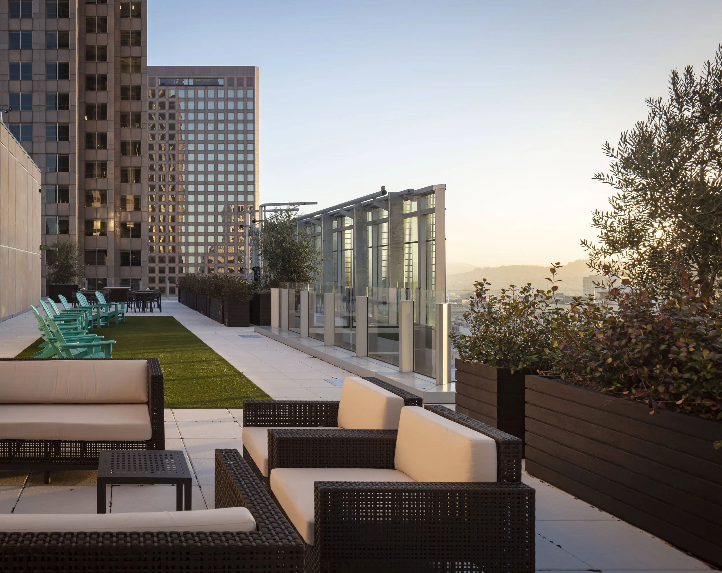 Rooftop terrace with lounge chairs, armchairs, and tables, surrounded by potted plants, with city buildings in the background at sunset.