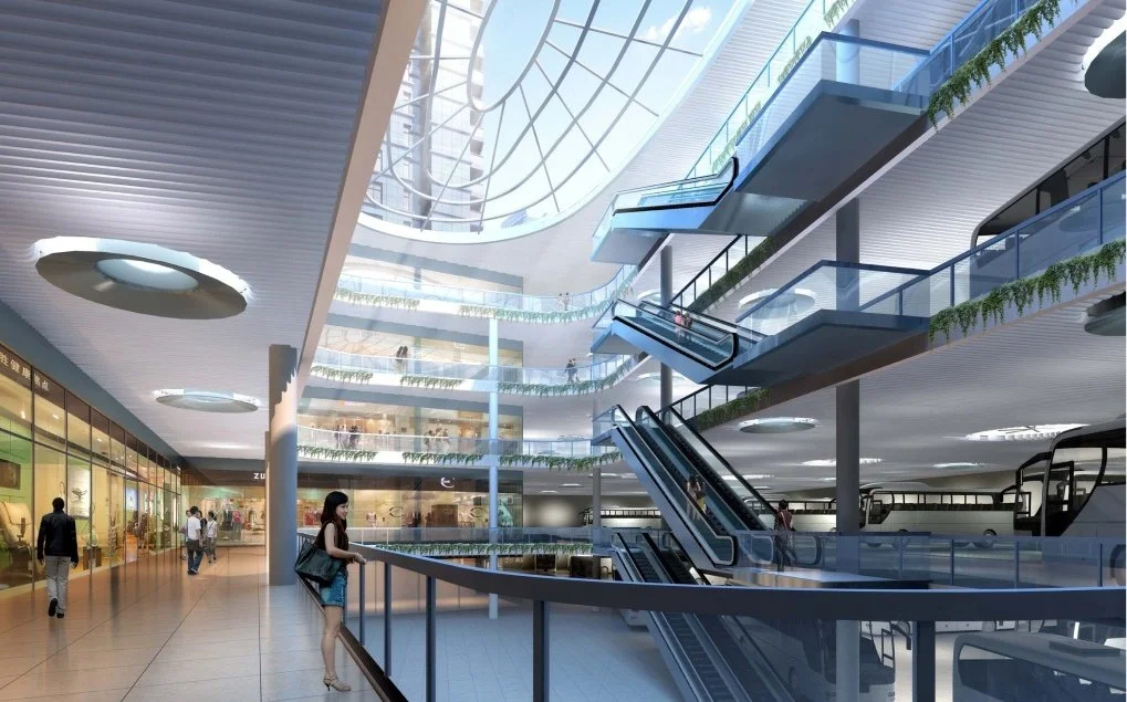 Interior of a modern shopping mall with multiple floors, glass railings, escalators, and a glass ceiling allowing natural light.