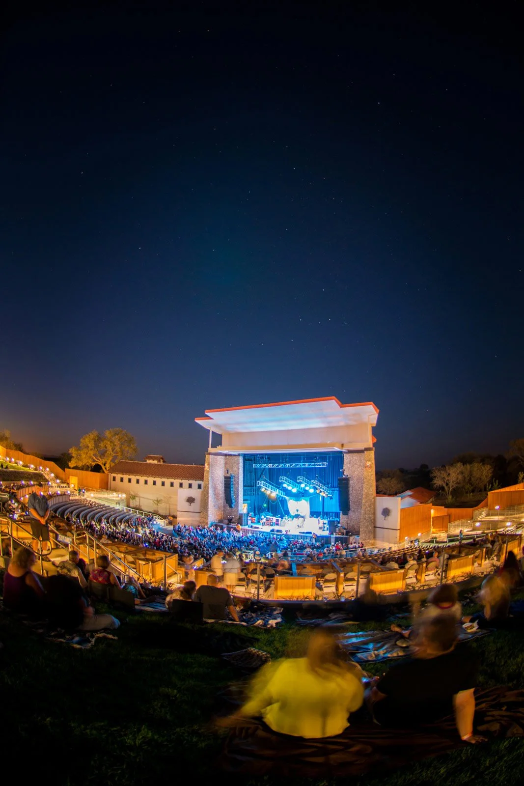 Outdoor amphitheater at night with people watching a live concert on stage, under a starry sky, with surrounding trees and seating area.