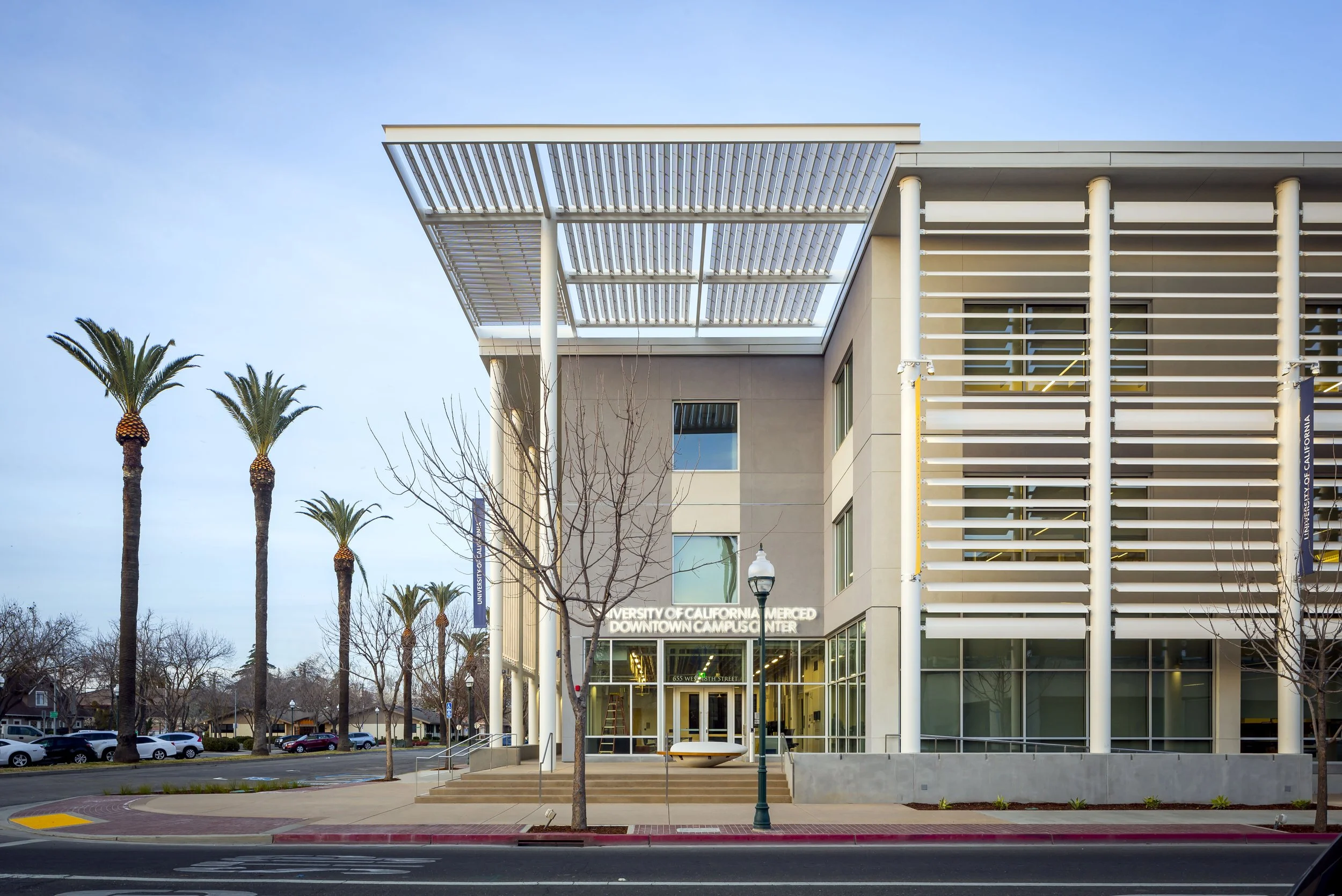 Front view of the University of California Merced Downtown Campus Center, with stairs, trees, street, and palm trees in the background.