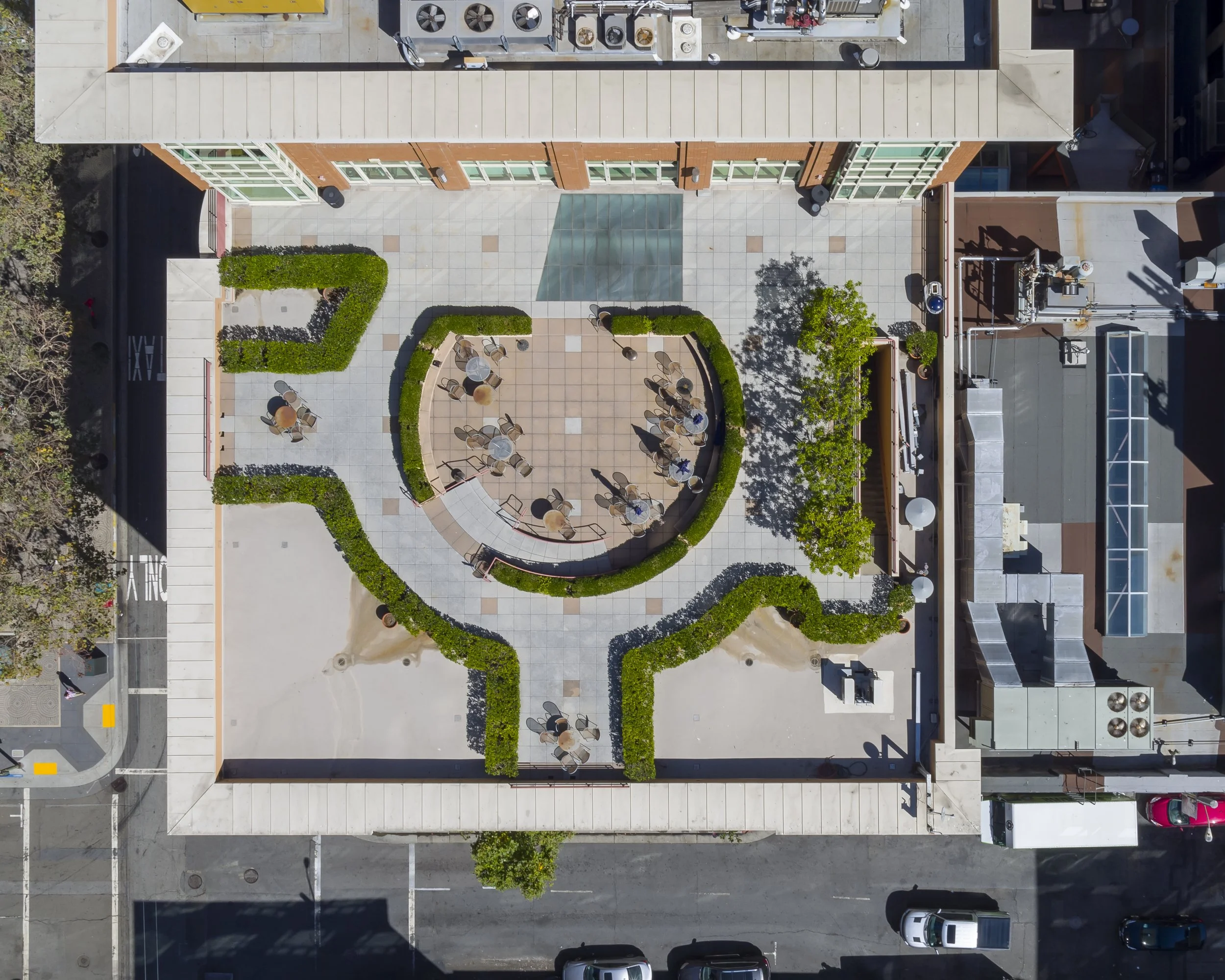 An aerial view of an outdoor rooftop patio with a semi-circular seating area surrounded by green hedges, tables and chairs, some with people, and nearby trees and buildings.