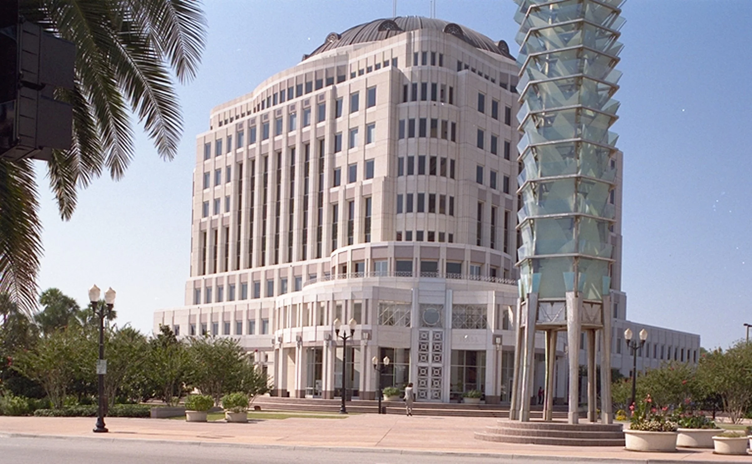 A tall modern building with a rounded top and a unique glass spiral tower next to it, set against a blue sky with some trees and street lamps in the foreground.