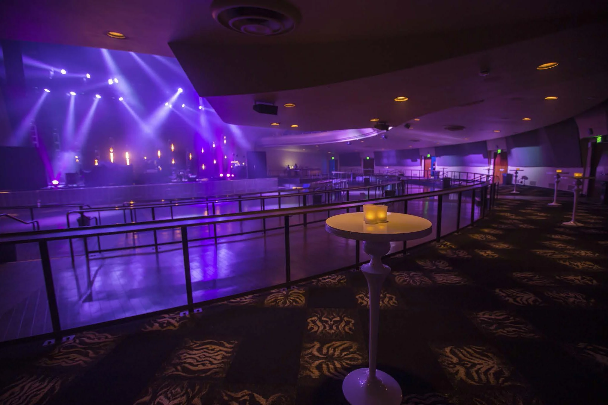 Empty indoor event venue with purple lighting, a stage with spotlights, and a dance floor enclosed by a black railing. Candles on tables provide warm light.