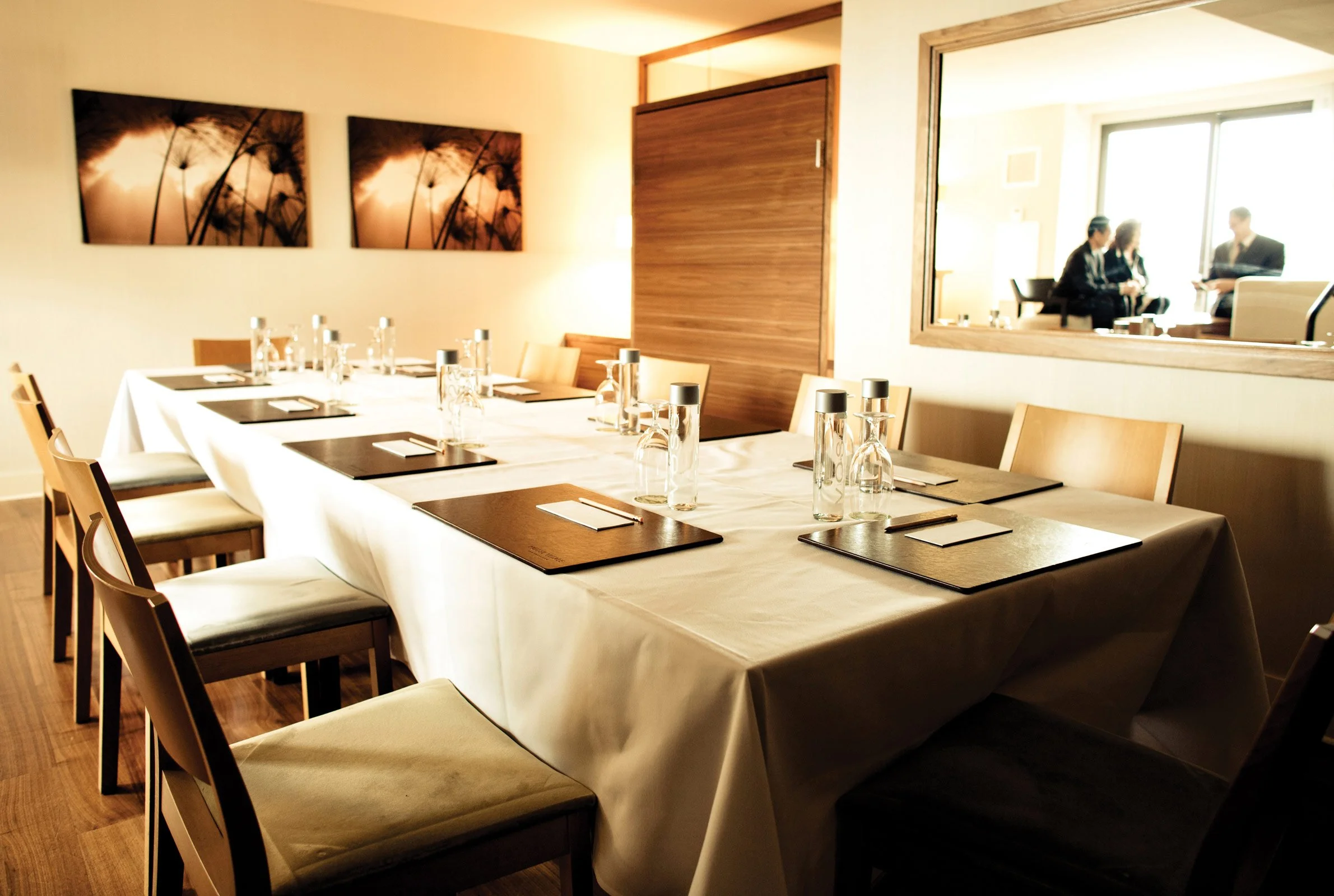 A private dining room with a long table set with water bottles, glasses, brown placemats, and napkins, with artwork on the wall and a view into an adjoining room with people sitting and chatting.
