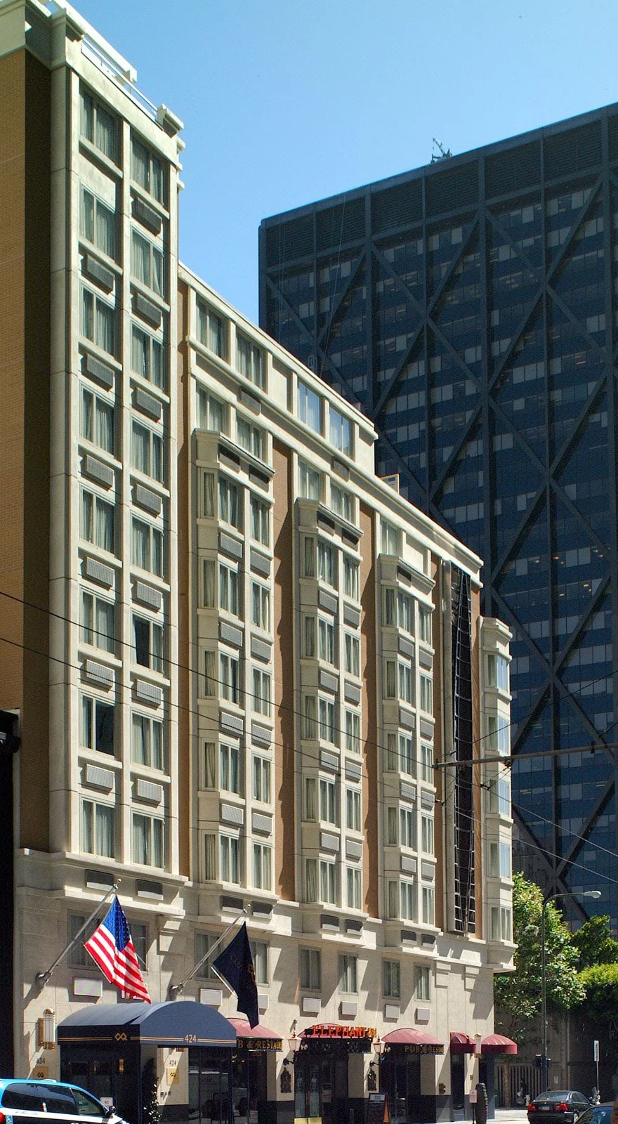 City street with mid-rise and high-rise buildings, American flags in front of a hotel or restaurant, streetlights, cars, and overhead wires under a clear blue sky.
