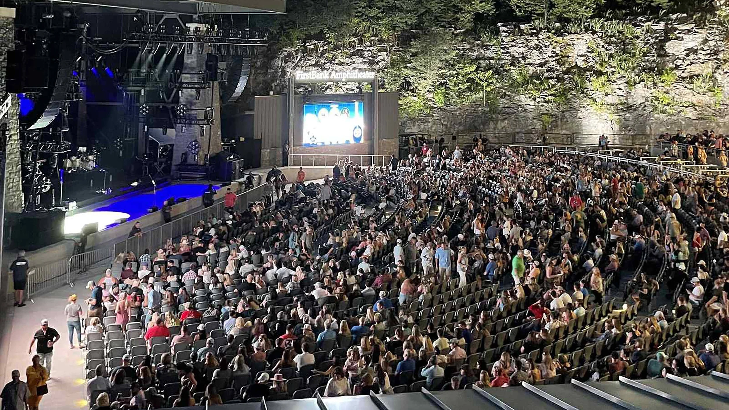A large outdoor amphitheater filled with seated audience members waiting for a performance, with a stage on the left and a large screen at the back of the stage.