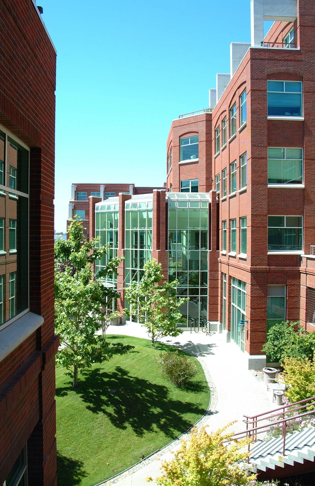 Urban courtyard surrounded by red brick buildings with large glass windows and doors, green trees, and a well-maintained lawn, under a clear blue sky.
