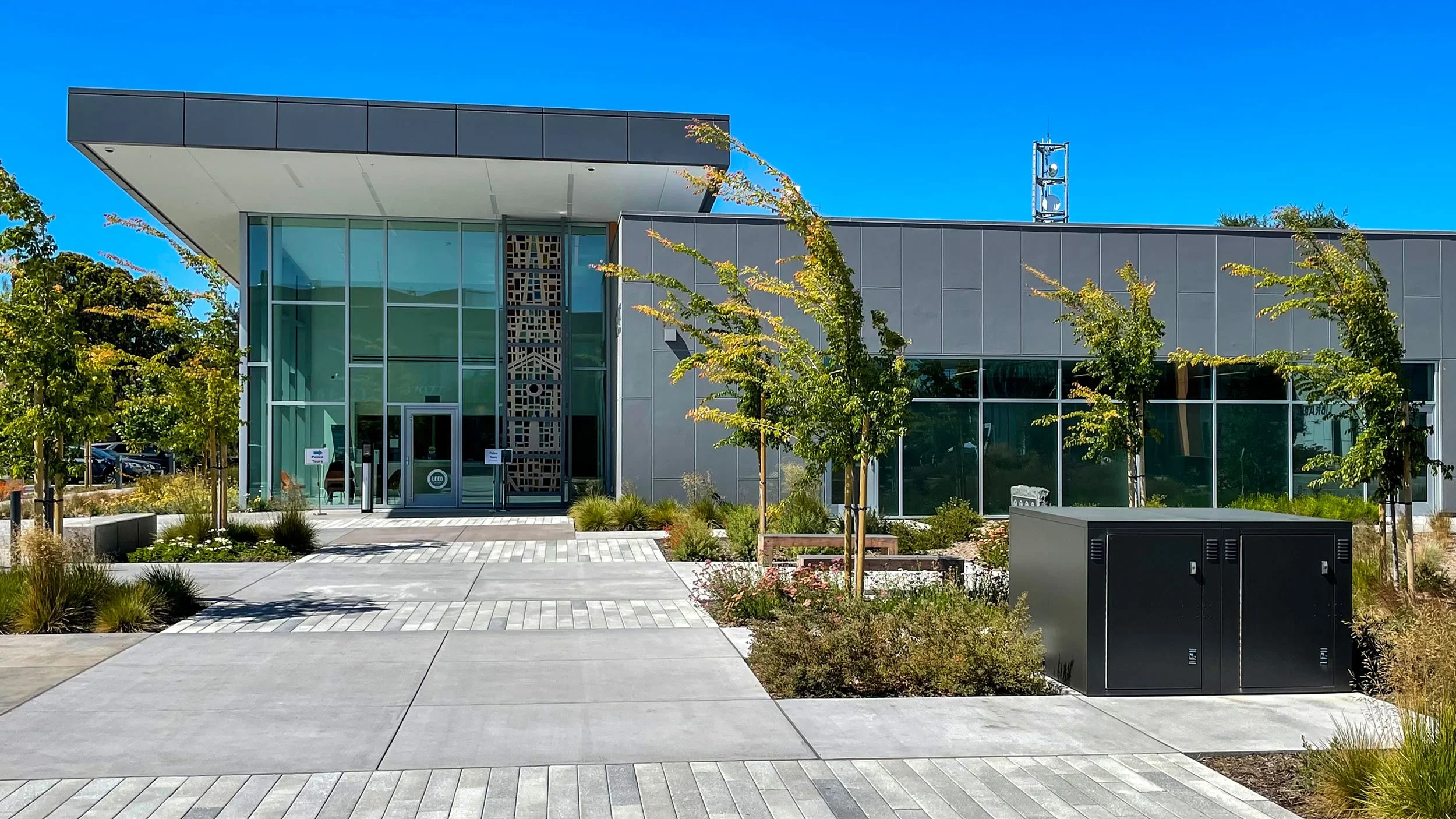 Modern building with large glass windows, a landscaped front with trees and shrubs, and a black utility box near the sidewalk. Clear blue sky overhead.