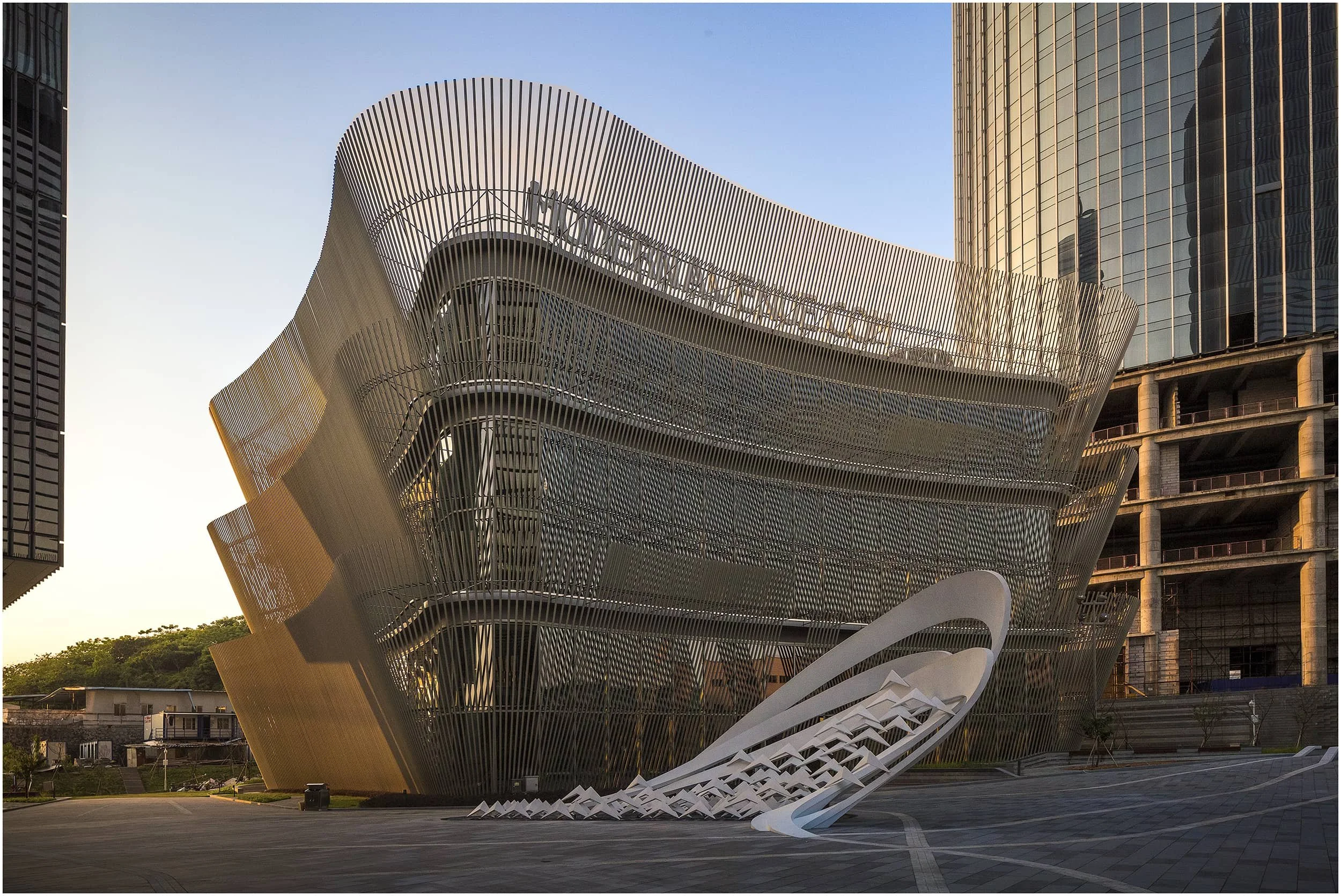 Modern architectural building with curving metal facade and a large, sleek white sculpture in front, set in an urban environment during sunset.