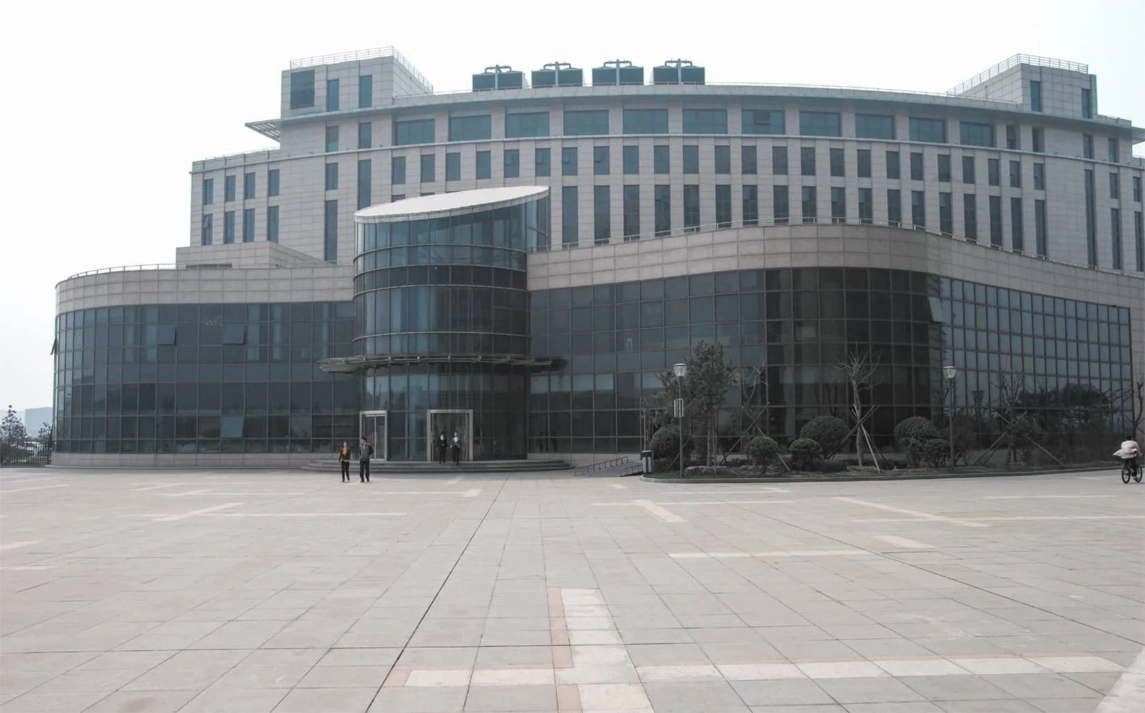 Modern multi-story building with glass windows and a curved architectural design, surrounded by an open paved area with a few individuals and a cyclist.