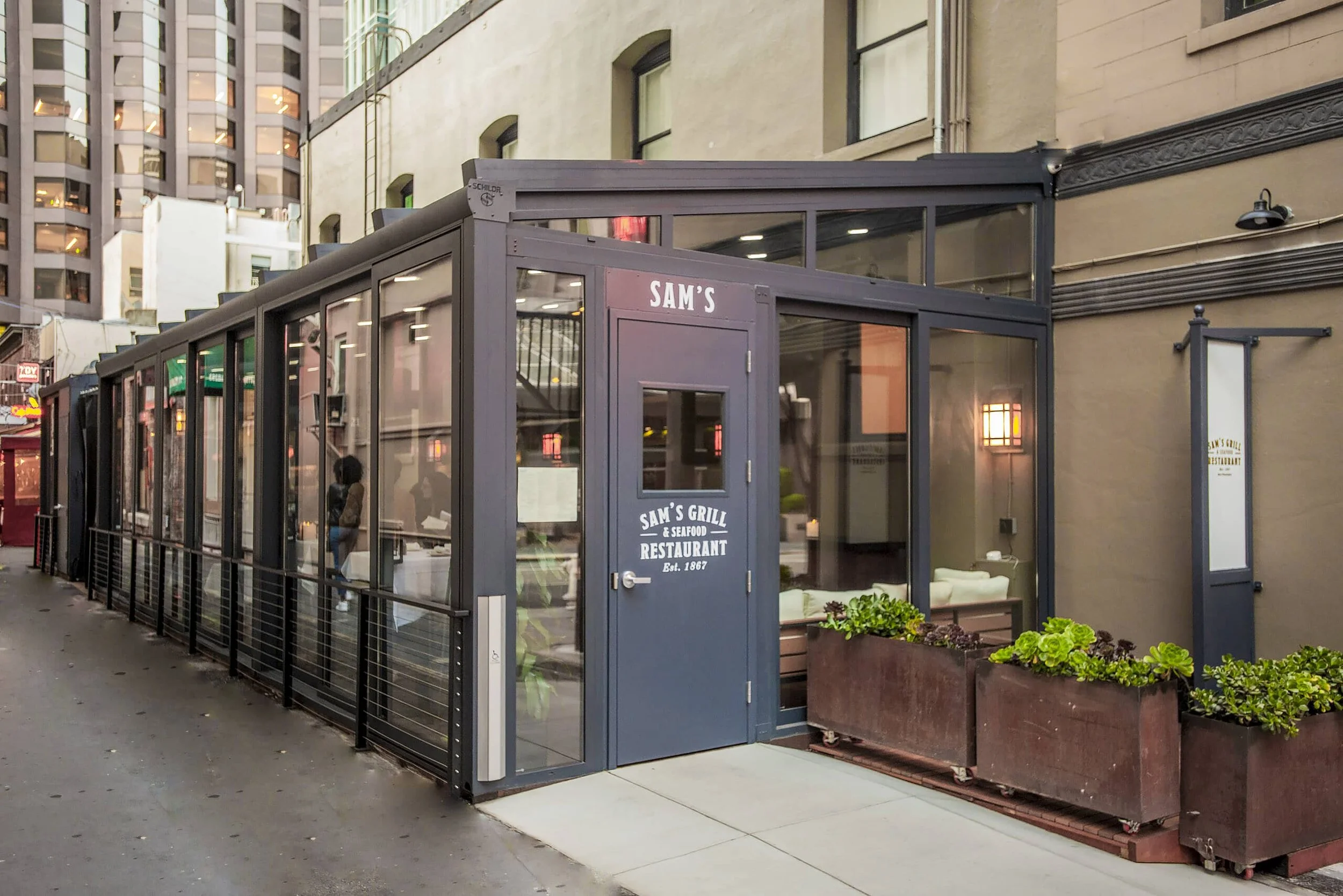 Exterior view of Sam's Grill & Seafood Restaurant in a narrow building with glass walls and a blue door, located on a city street.