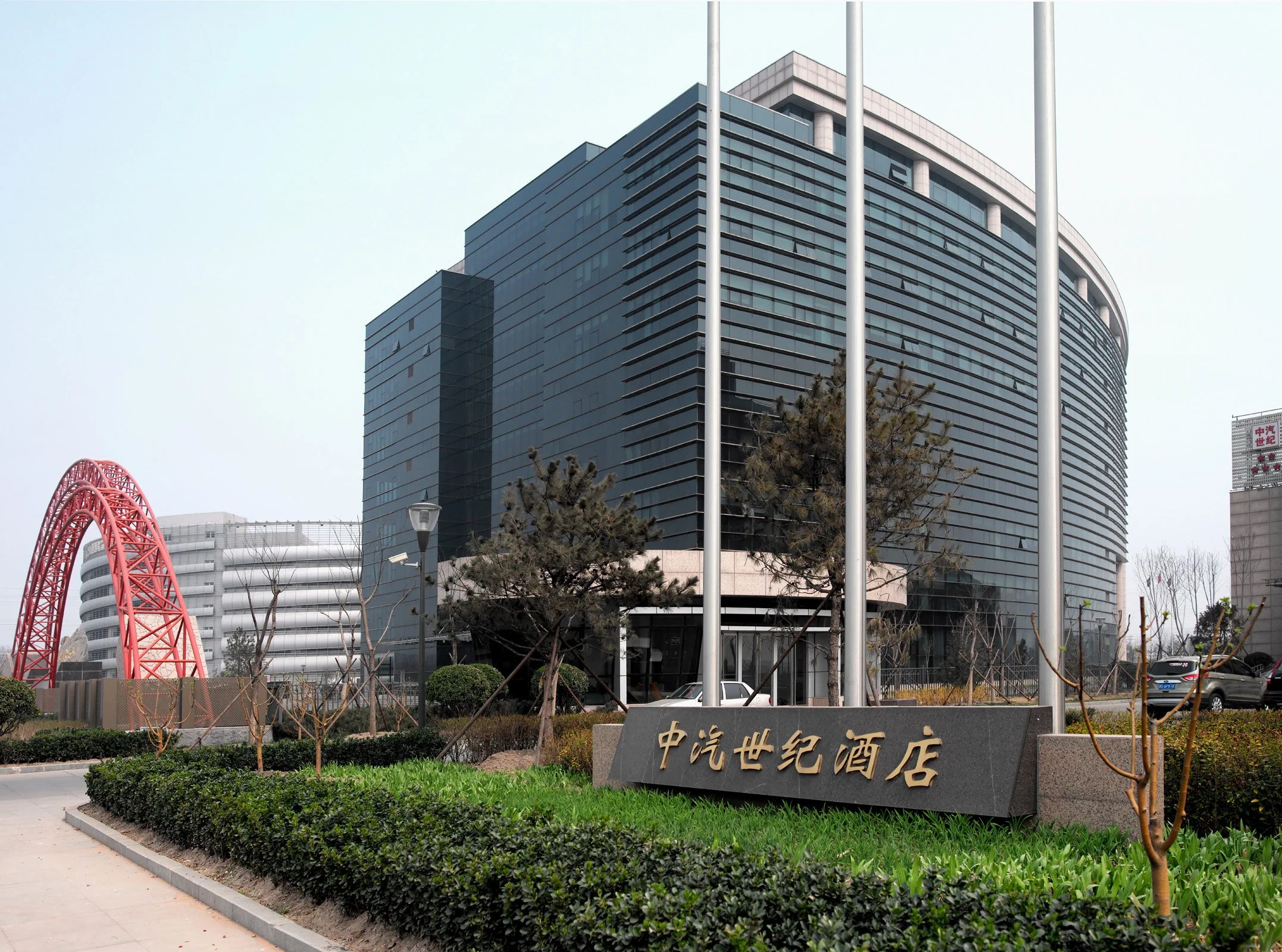 Modern office building with glass facade, a red bridge in the background, and a variety of landscaped trees and shrubs in the foreground, including a sign with Chinese characters.