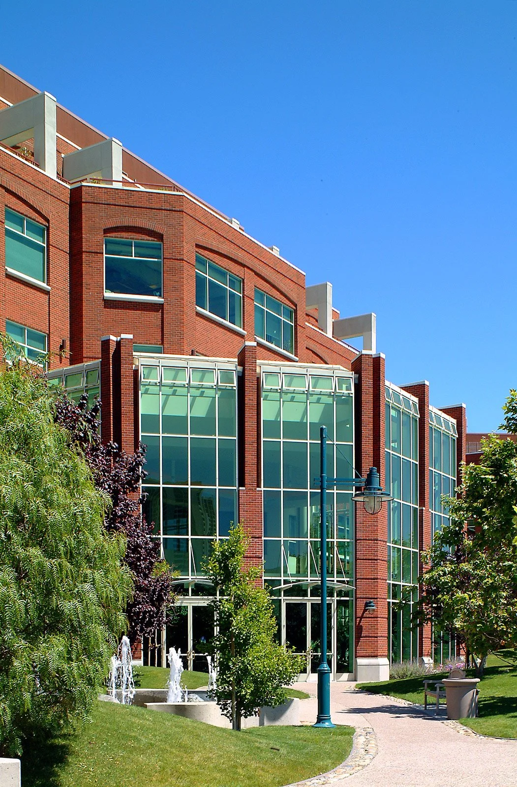 Modern brick building with large glass windows, surrounded by green trees, a pathway, benches, and fountain in a park setting under a clear blue sky.