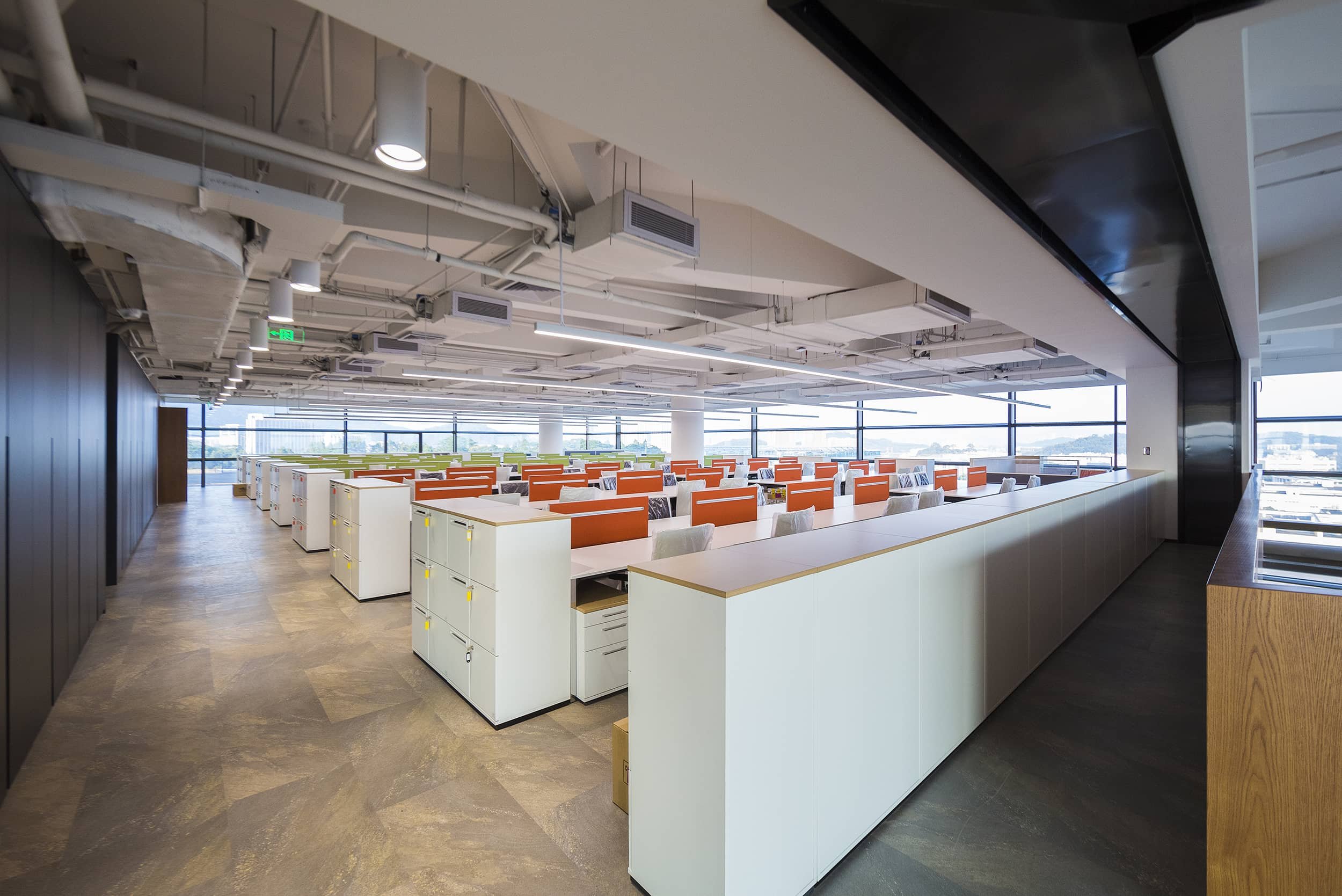 Empty modern office space with rows of white desks and orange dividers, large windows with city view, exposed ceiling with pipes and lighting.