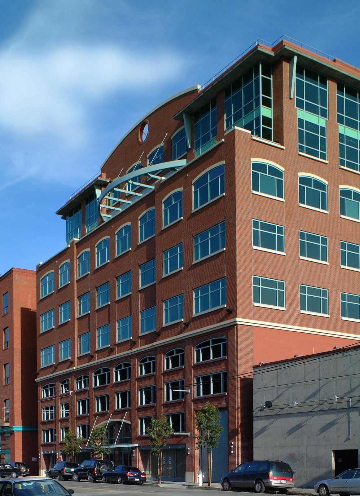 A tall multi-story brick building with many windows and a modern architectural design, with a partial street view in front and a partly cloudy sky above.