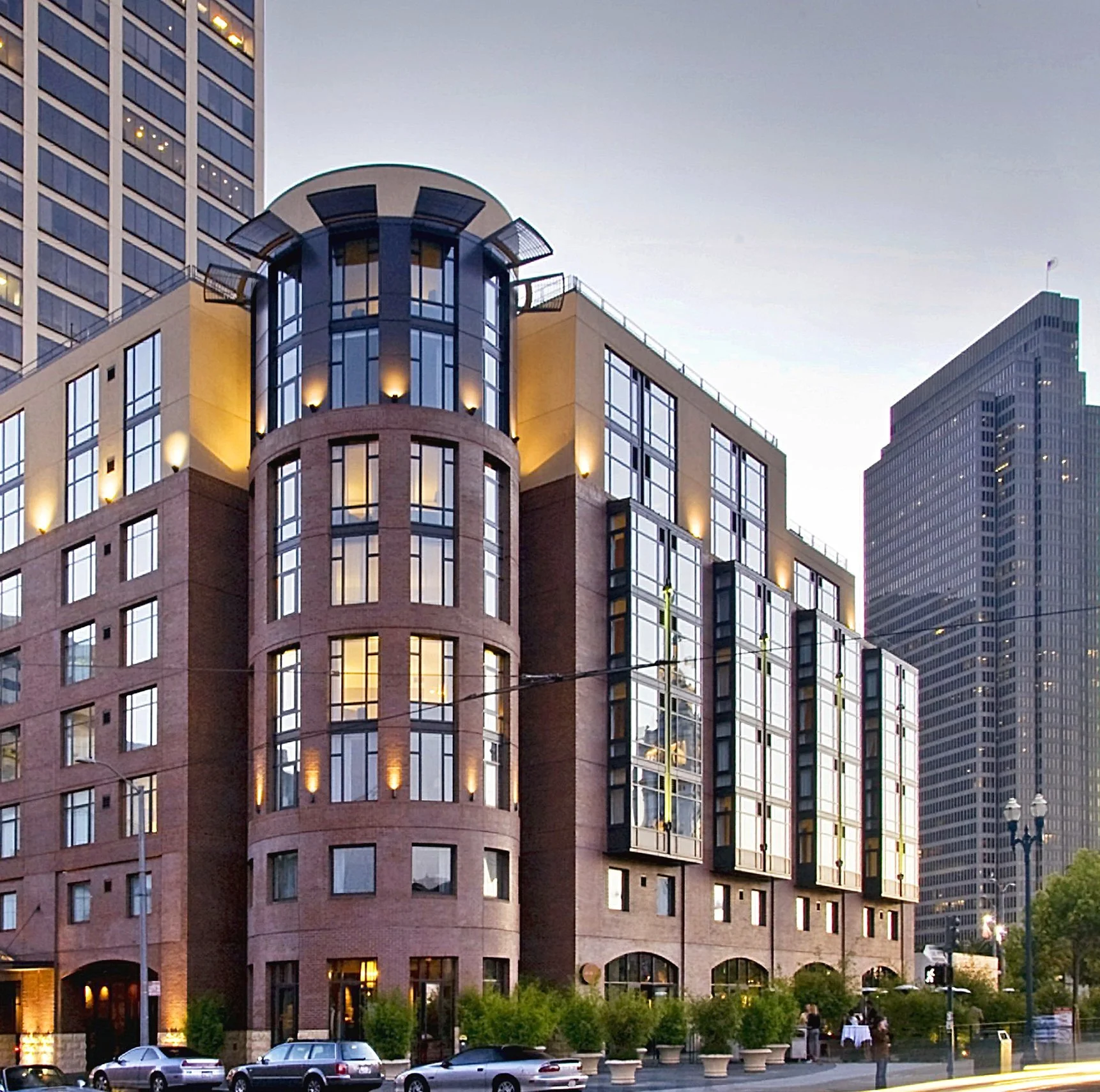Modern multi-story building with large glass windows, brick and metal exterior, and trees in front, set in an urban environment during evening.