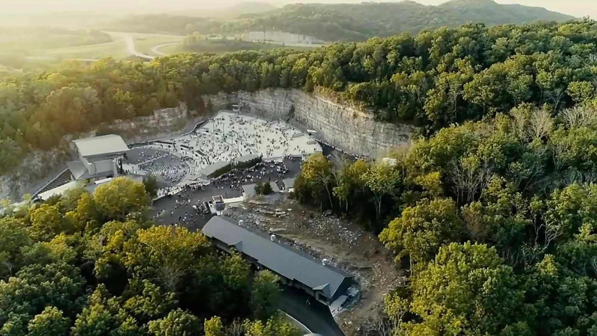 An aerial view of an outdoor amphitheater built into a hillside surrounded by dense green forest with some construction or damage visible at the front of the stage area.
