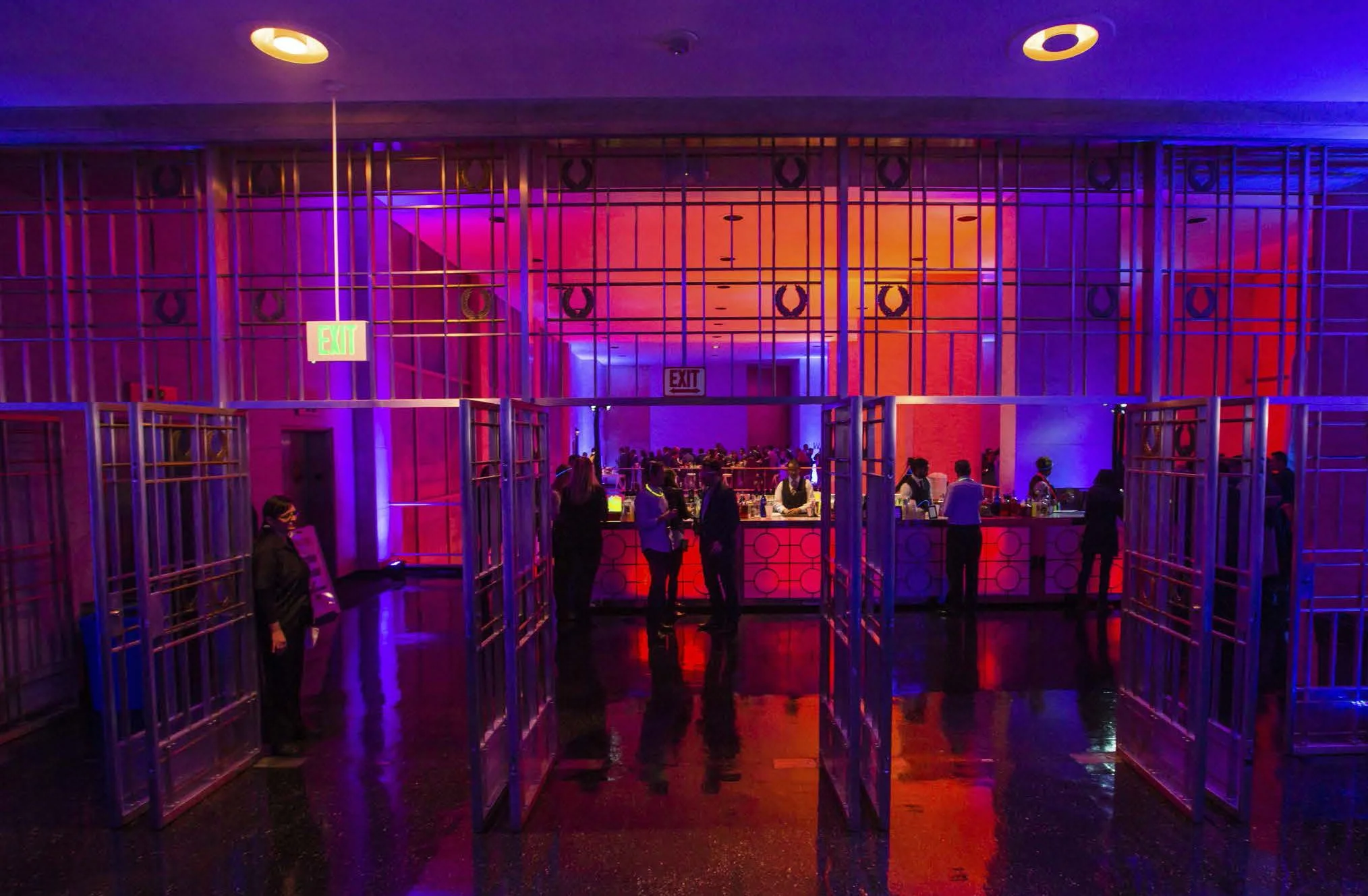 Indoor bar area with purple, pink, and blue lighting, featuring a metal gate design and a bar counter with bartenders and customers.