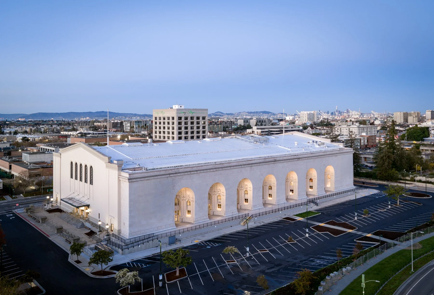 An aerial view of a white church with large arched windows and an empty parking lot in front, with a cityscape and distant hills in the background during early evening.