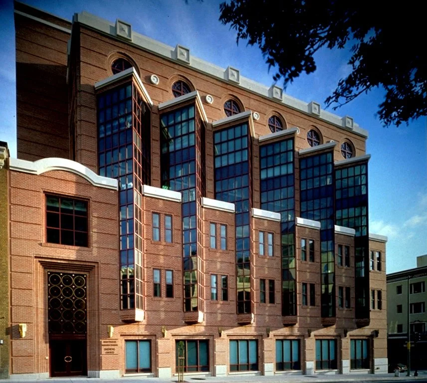 A multi-story brick building with large blue-tinted glass windows and decorative architectural details, set against a blue sky with some tree branches in the foreground.