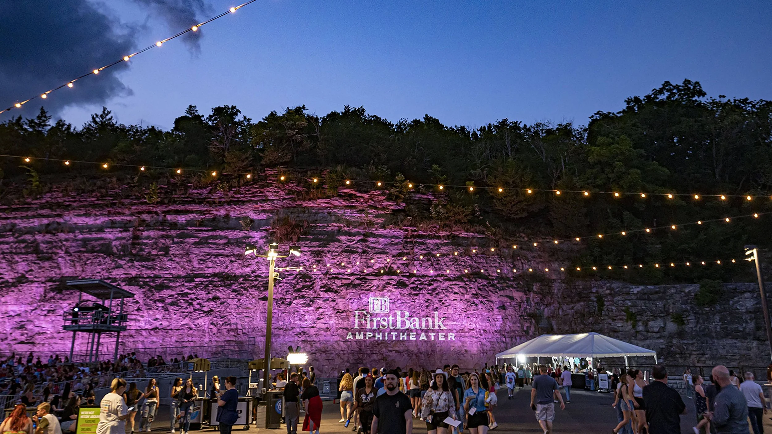 Crowd of people gathered outside the First Bank Amphitheater at dusk, with string lights overhead and a rocky cliff face illuminated in pink in the background.