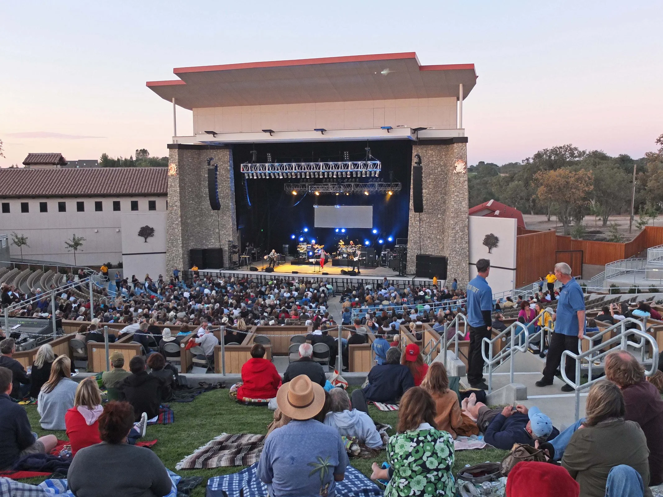 An outdoor concert at dusk with a stage featuring musicians and colorful lights, overlooking an audience seated on the grass and in tiered seating, enjoying the performance.