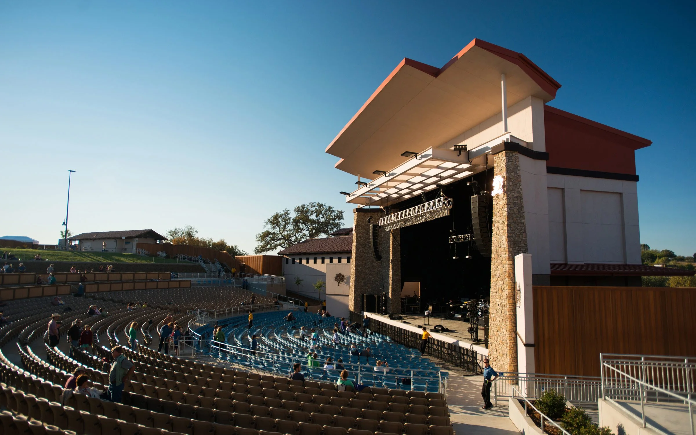 Open outdoor amphitheater with a large stage and tiered seating, some people walking and sitting, clear sky