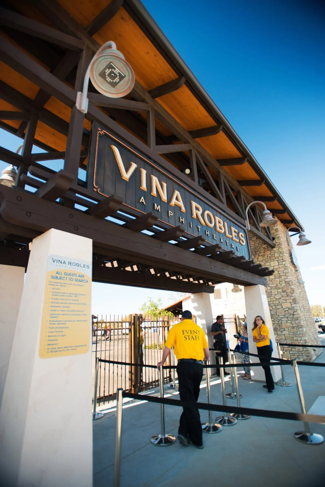 The entrance of the Vina Robles Amphitheatre with a large sign, event staff in yellow shirts, and security checks at the gate against a clear blue sky.