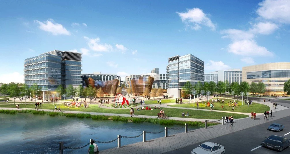 A modern city park with green lawns, walking paths, and a pond in the foreground. There are people walking, biking, and relaxing, with several contemporary office buildings and sculptures in the background.