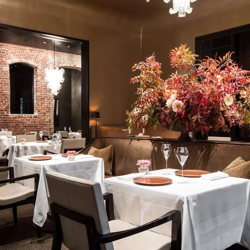 Elegant restaurant with white tablecloths, floral centerpieces, and a large floral arrangement with pink, red, and white flowers. The decor includes exposed brick walls, modern chairs, and a chandelier.