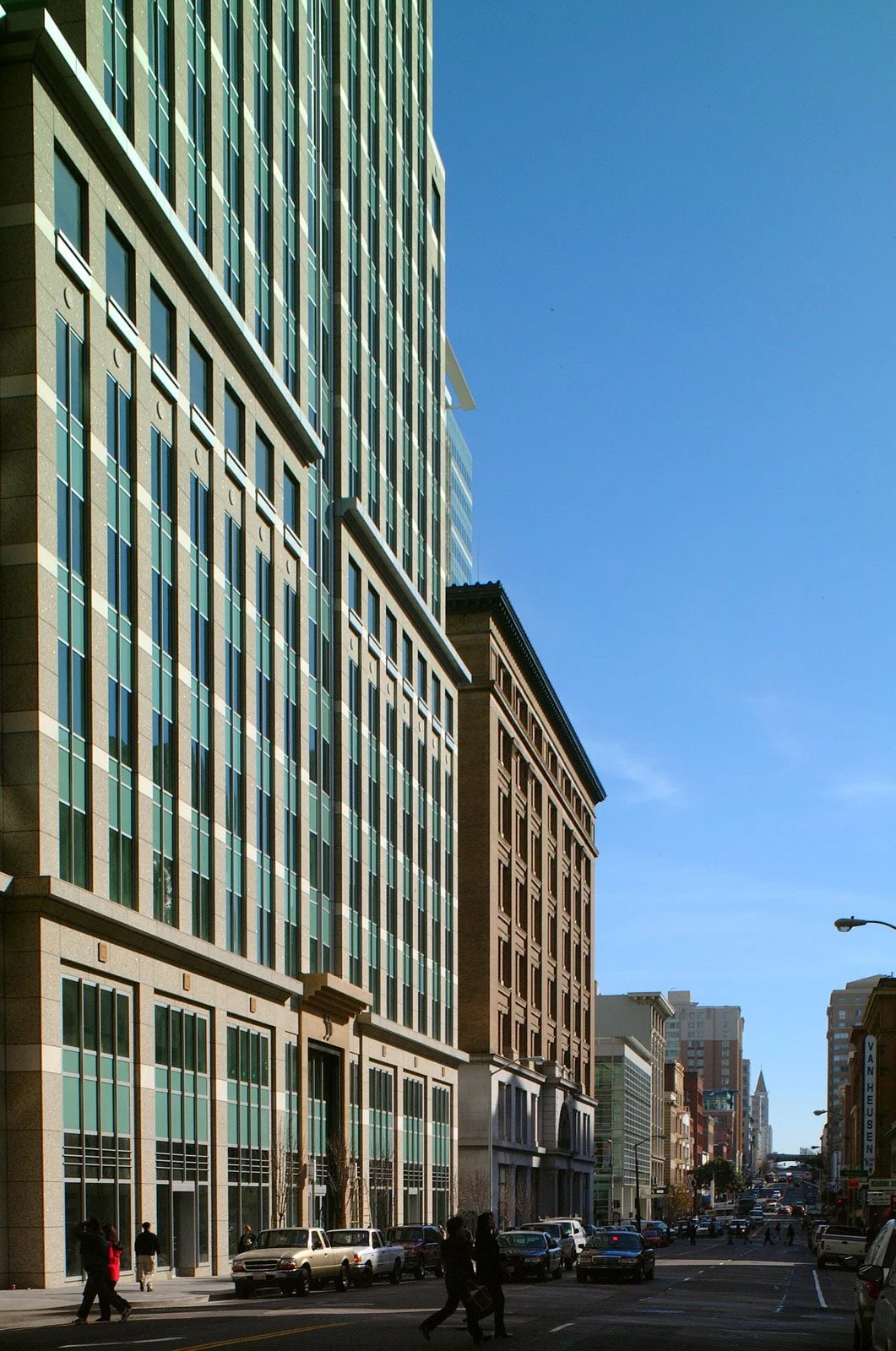 City street scene with tall modern glass buildings on the left and older brick buildings on the right, pedestrians walking, and cars parked along the street on a sunny day with a clear blue sky.