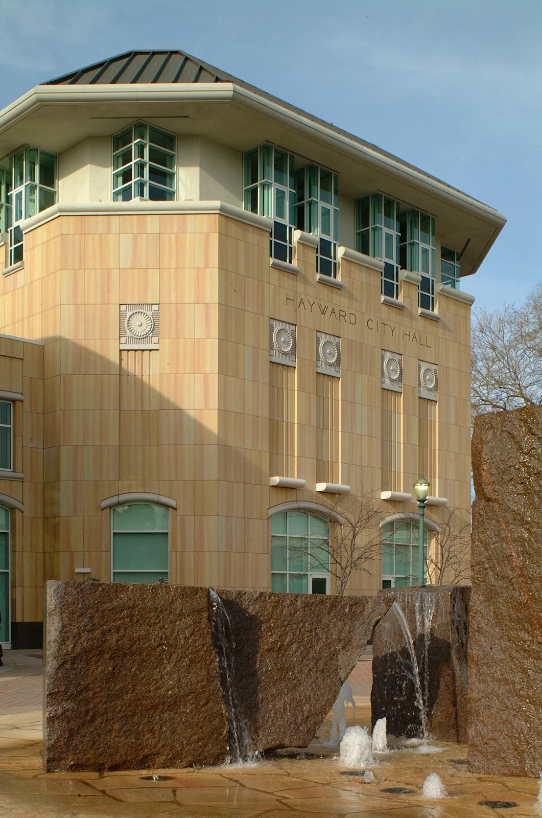 Hayward City Hall building with a fountain in the foreground and a lamp post, under a clear blue sky.