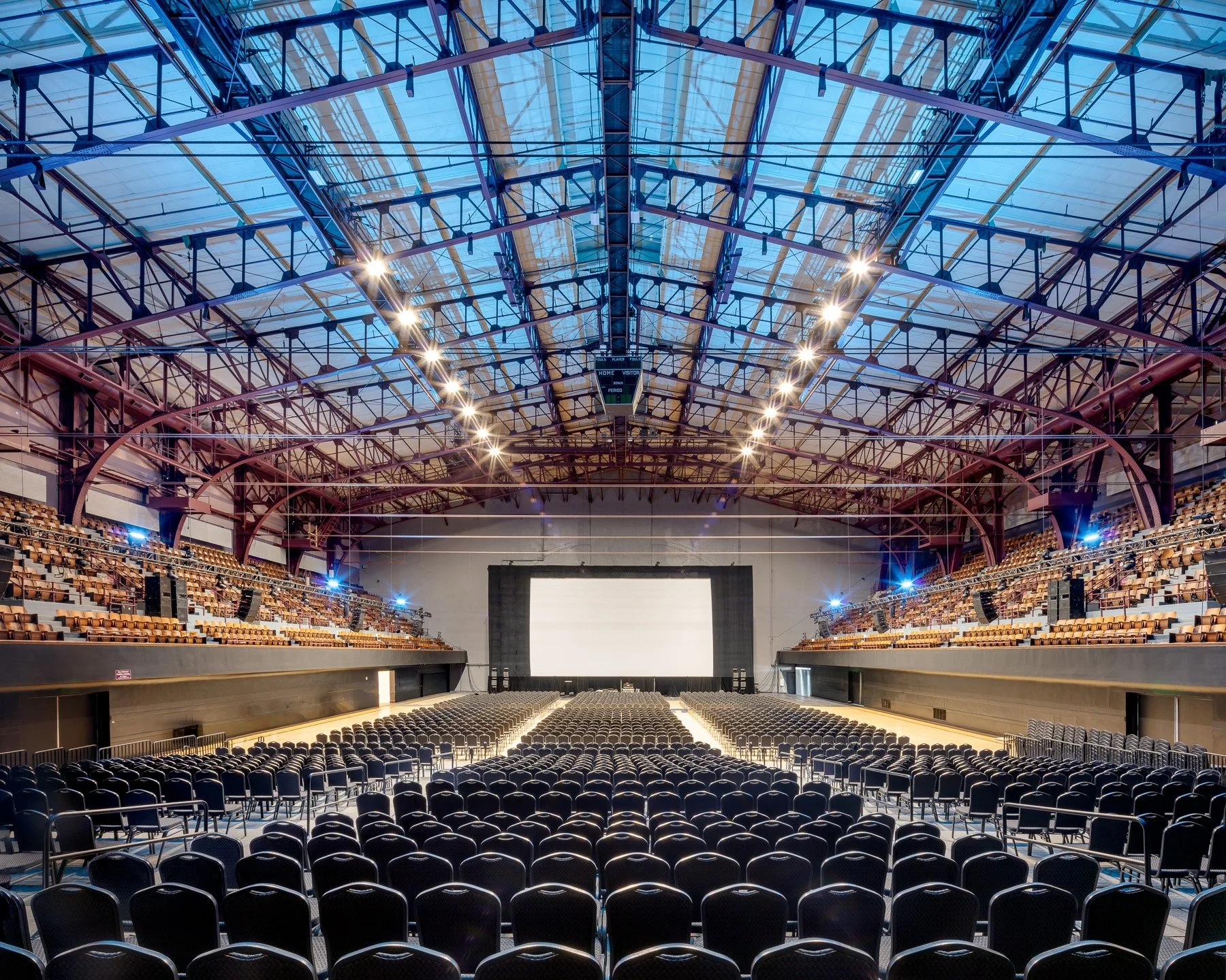 Empty indoor sports arena with rows of black chairs facing a large screen or stage, glass ceiling, and metallic framework.