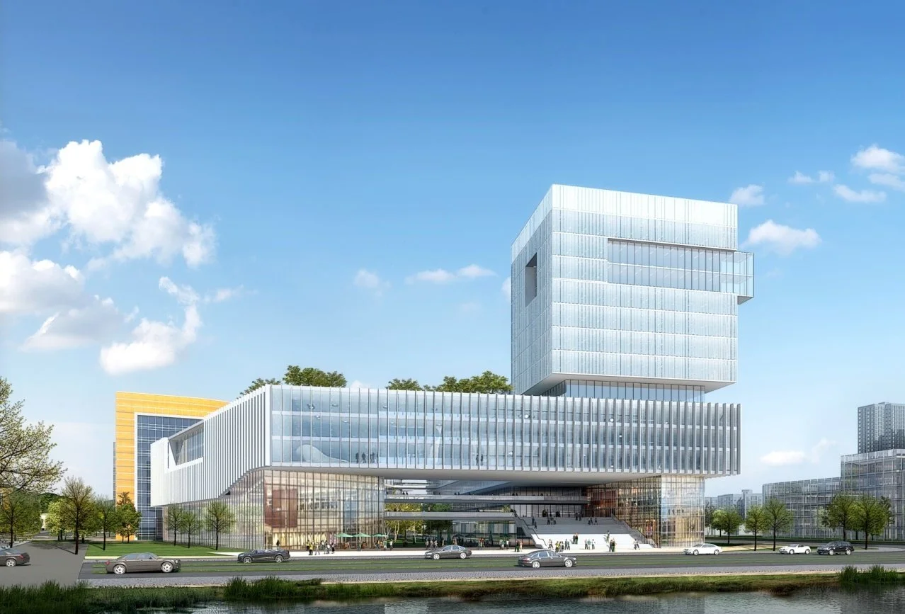 Modern multi-story glass building with a floating section, located near a waterway with cars and trees in the foreground.