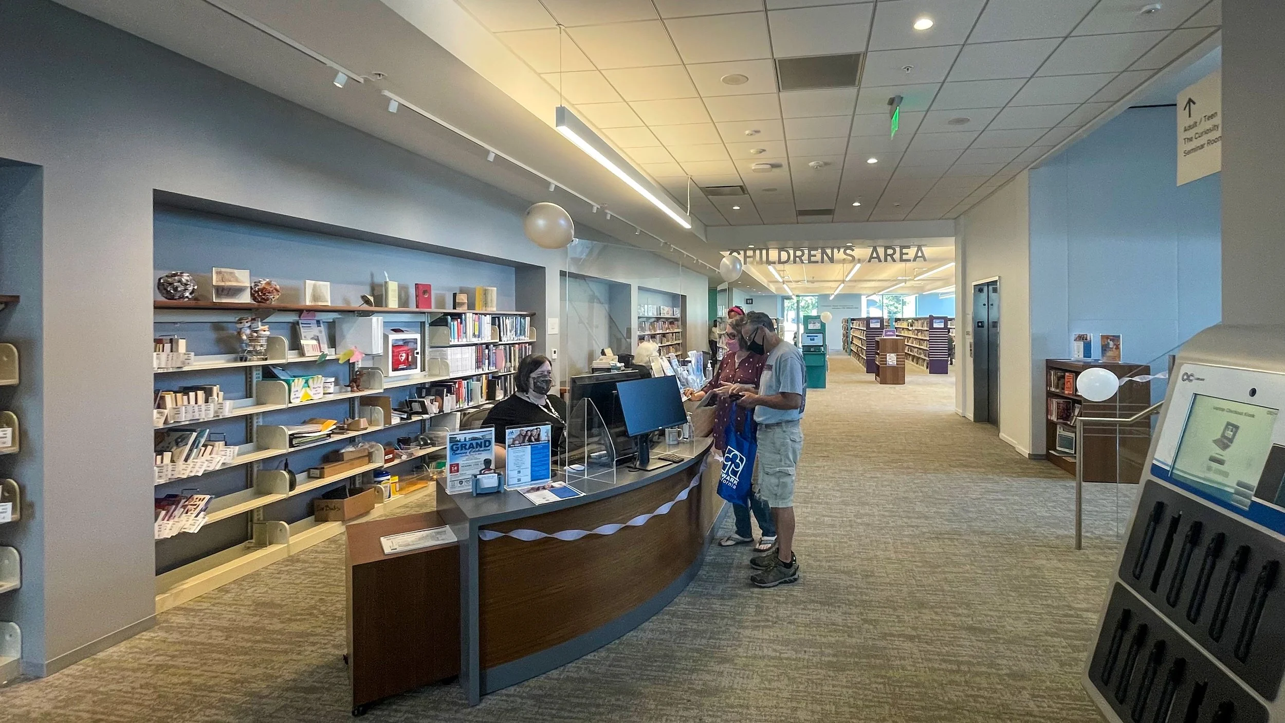 Inside a library, a receptionist at a curved desk is speaking with two visitors, all wearing masks. Behind the desk are shelves filled with books and decorative items. Further back, there are more bookshelves and a sign indicating the children's area