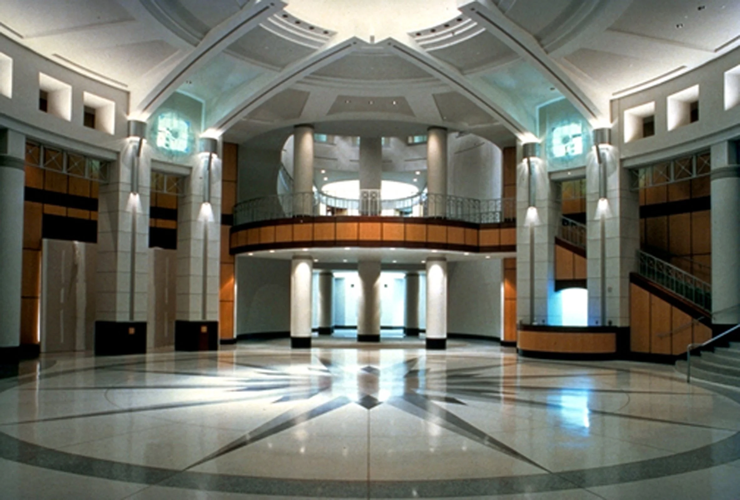 Interior view of a spacious, elegant lobby with marble floors, white columns, wooden accents, and a balcony with a metal railing, illuminated by soft lighting.