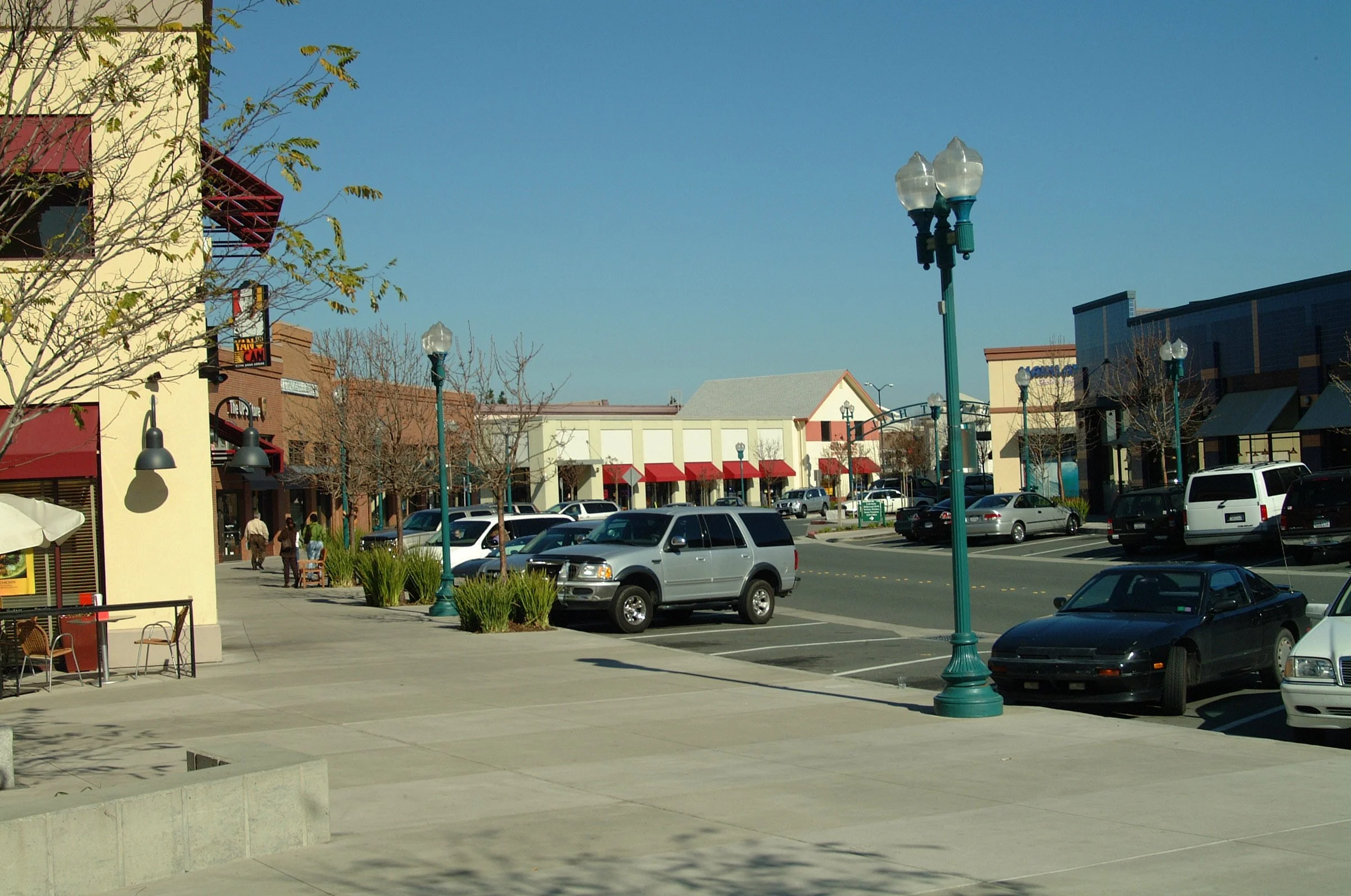 A shopping center parking lot with parked cars, street lamps, a sidewalk, and storefronts under a clear blue sky.